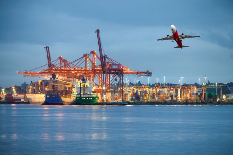 International cargo ship with logistics and containers cargo illumination gantry cranes and commercial airplane flying at import and export habour by the ocean