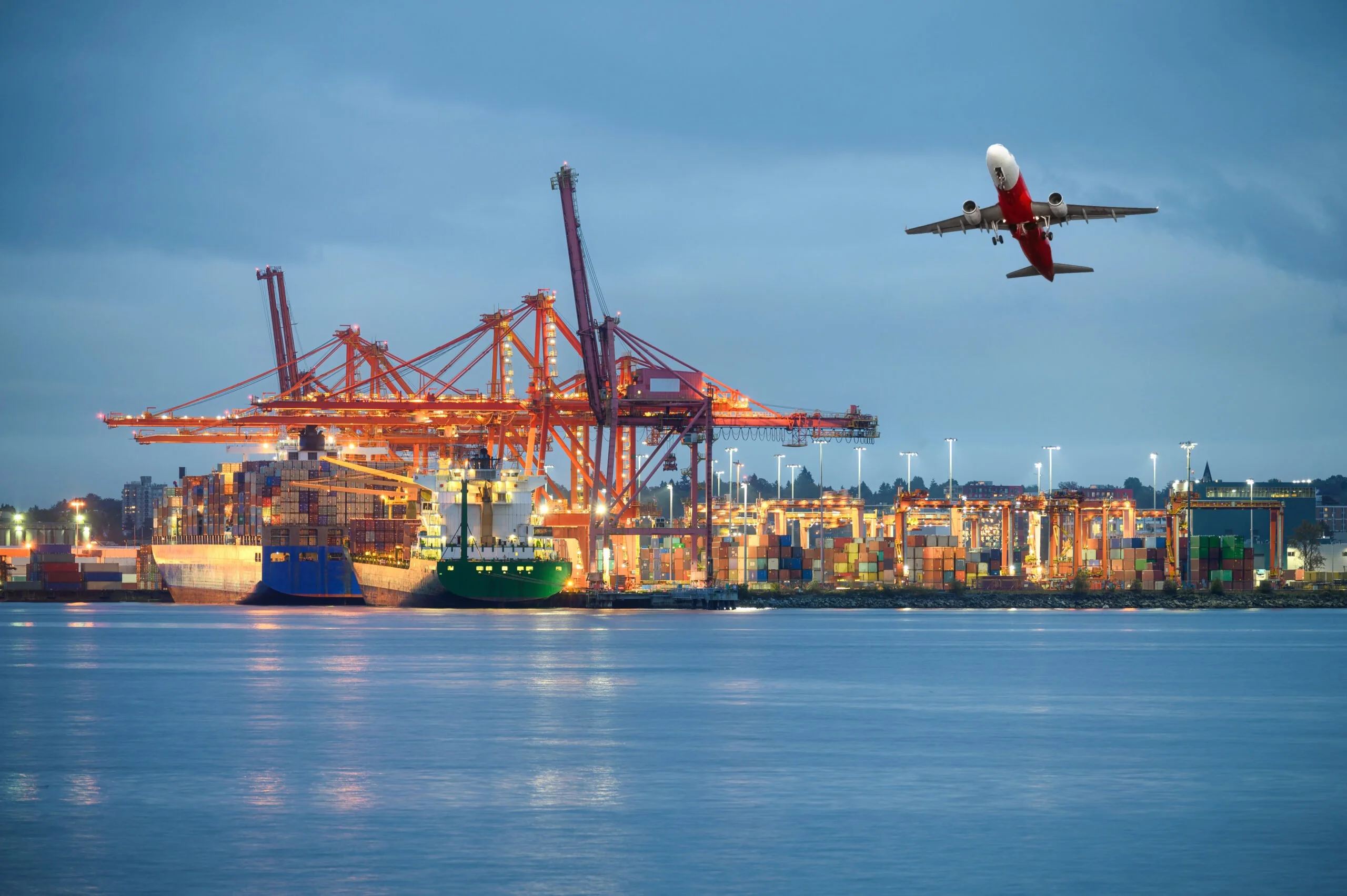International cargo ship with logistics and containers cargo illumination gantry cranes and commercial airplane flying at import and export habour by the ocean